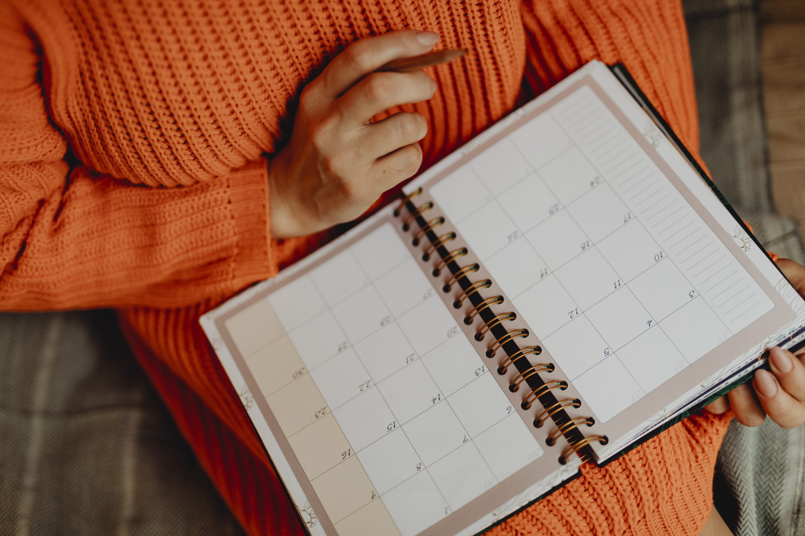 Pessoa com suéter laranja segurando um lápis e uma agenda aberta no calendário. O foco da imagem está na agenda