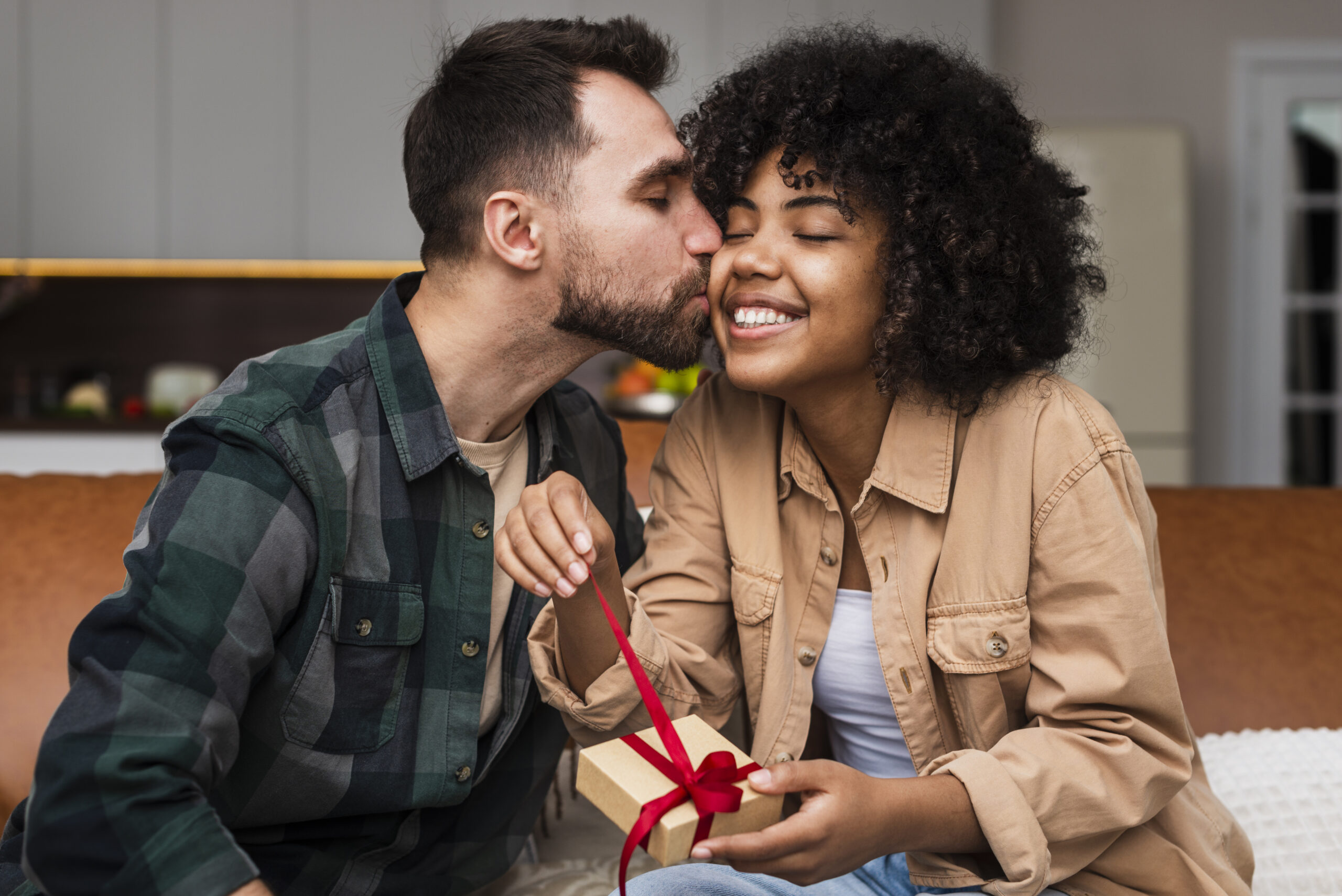 Homem branco de cabelos escuros e lisos dando um beijo na bochecha de uma mulher negra de cabelos escuros e enrolados enquanto ela abre uma caixa de presentes