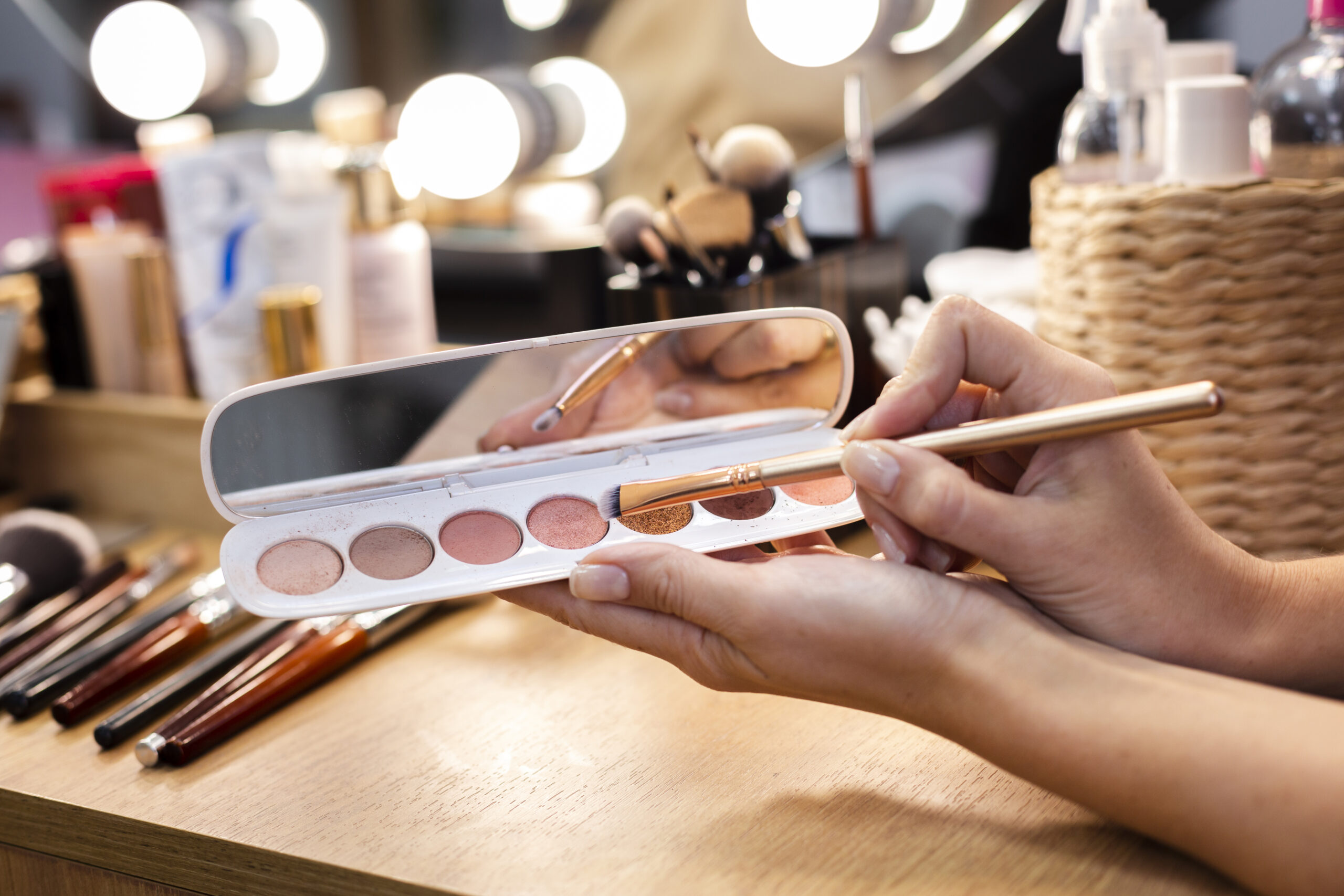 Mão segurando um kit de sombras de olho e um pincel de maquiagem em frente a uma bancada com produtos de maquiagem