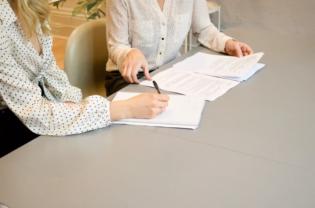 duas pessoas com camisas claras sentadas em uma cadeira, uma assina o papel da nota promissória enquanto a outra confere. 